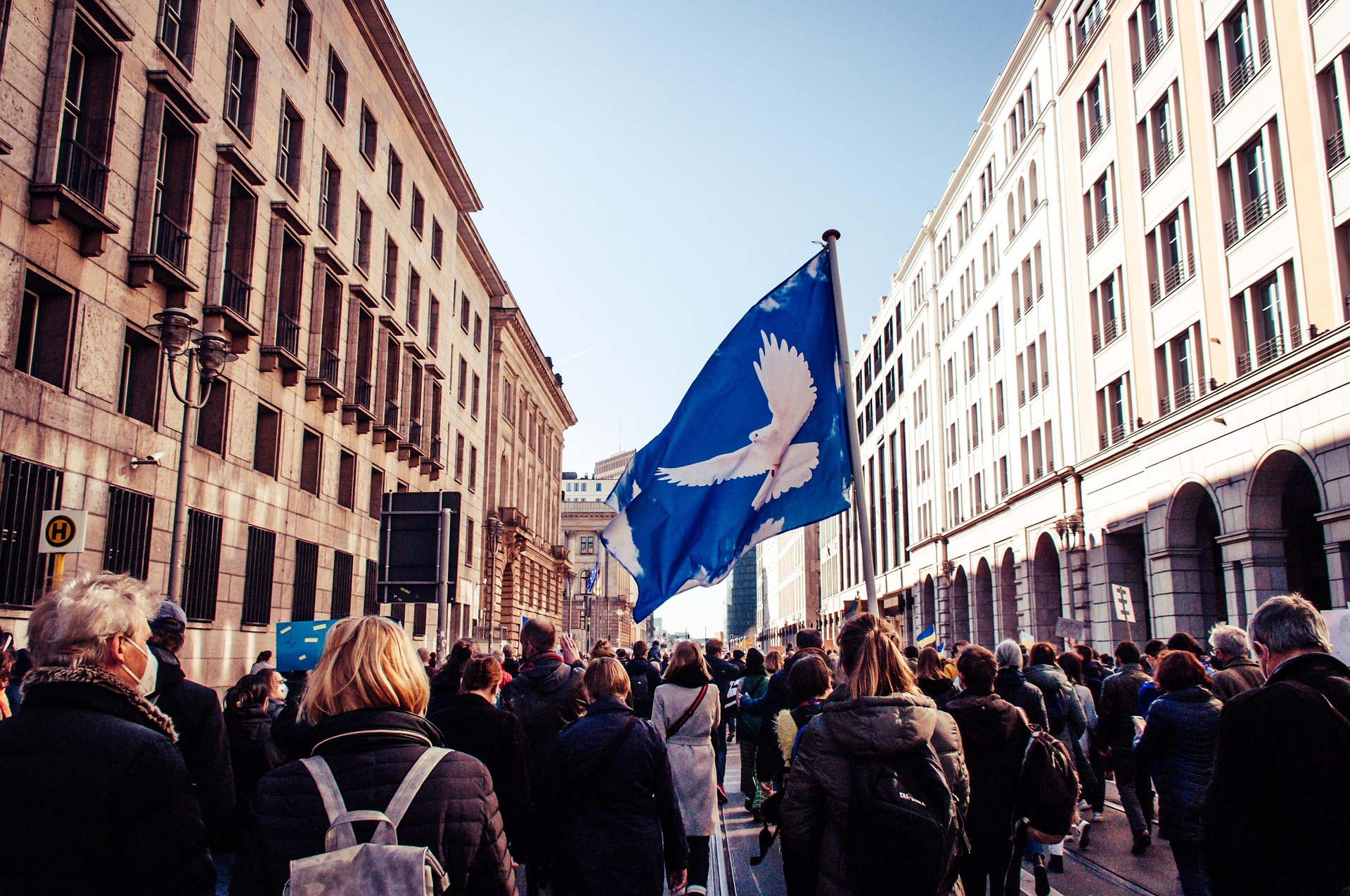A protest with a lot of people on a city street, with buildings on both sides. Most prominent of all is a Peace Flag held aloft, a white dove on a blue background.