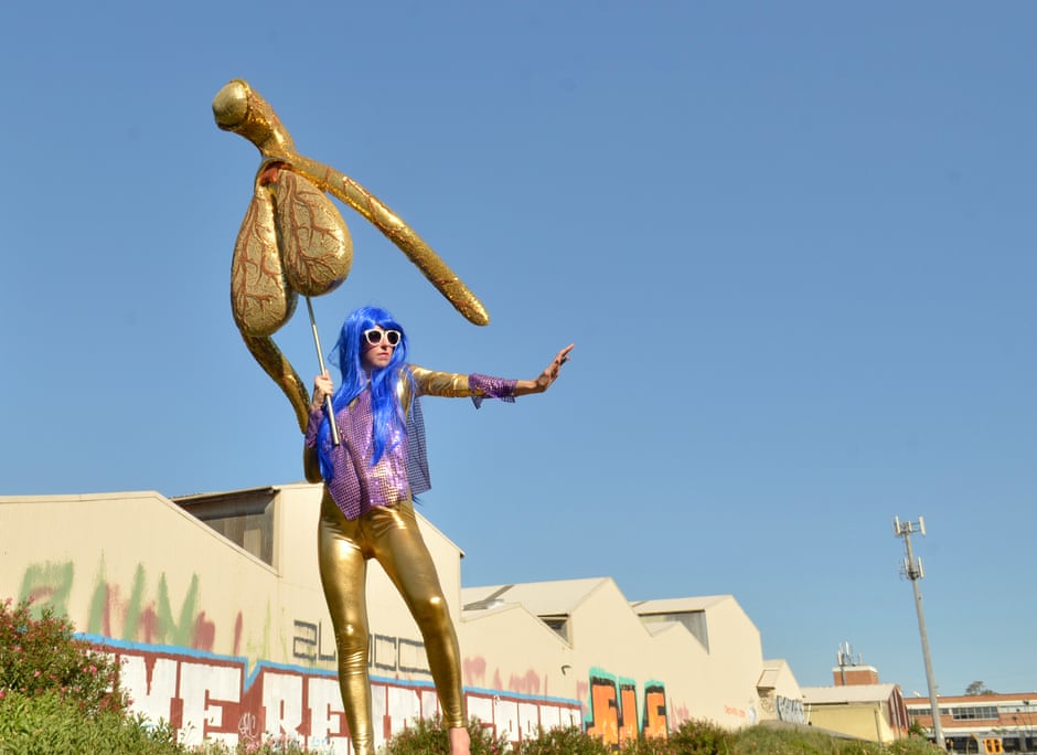Looking up at Alli Sebastian Wolf holding her 100-1 size gold clitoris, called 'Glitoris', with a background of sky