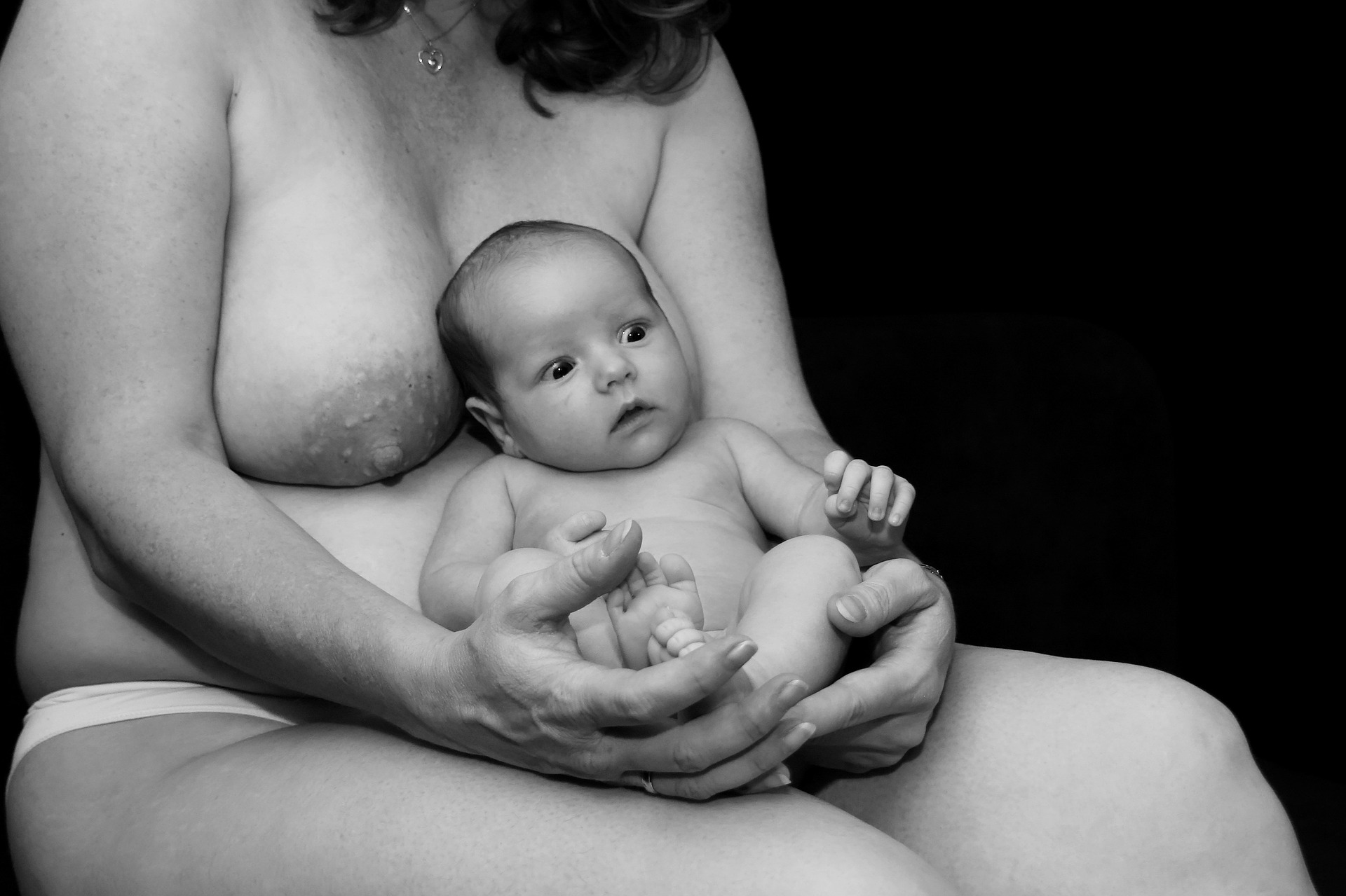 Surprised, even shocked, baby; or perhaps reflective; baby is seated and held warmly on the hips of the nude mother; baby's head rests between her breasts; mother's face not visible; B&W photo