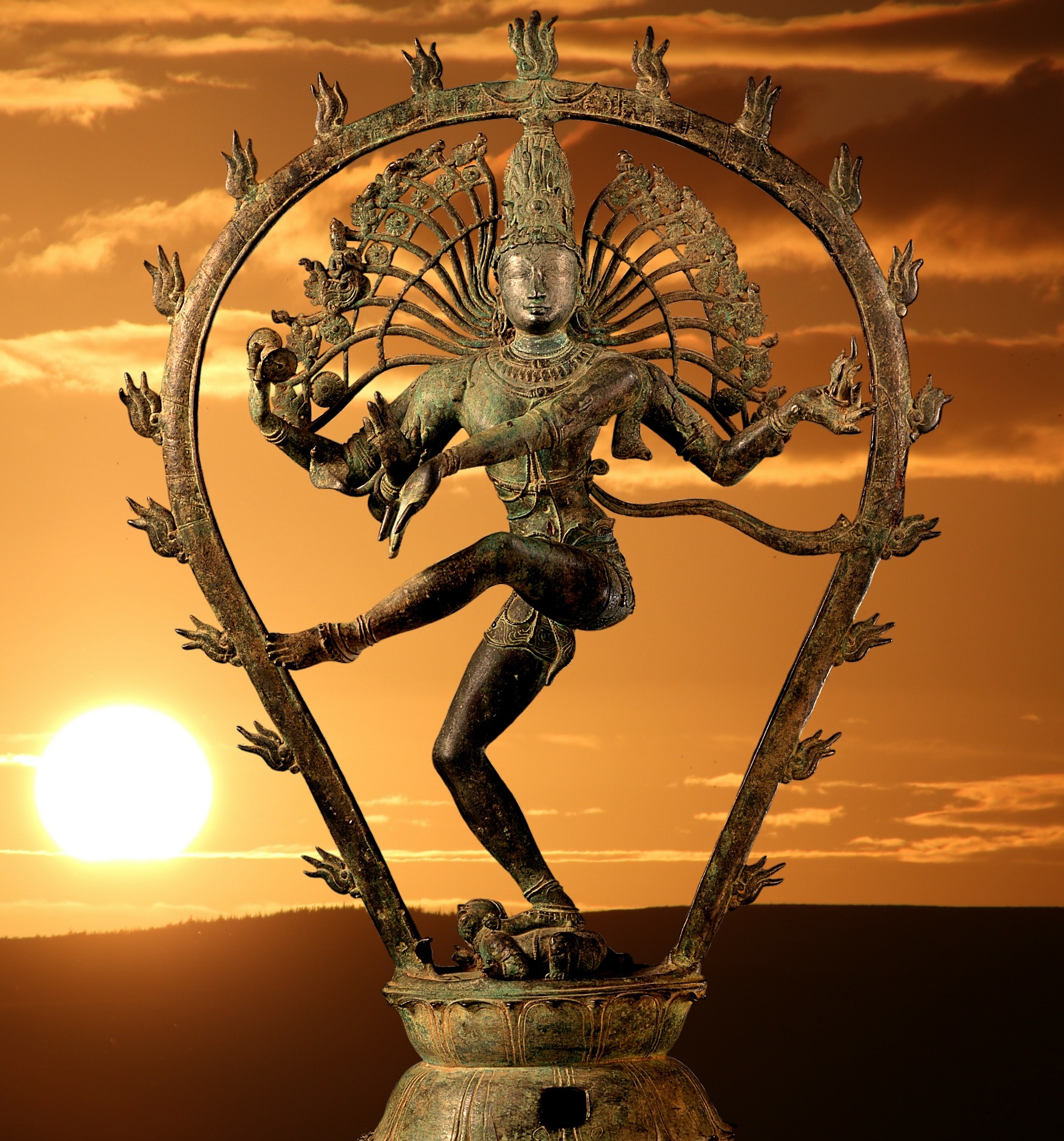 In the background, a beautiful glowing sun has just risen above a hill. In the foreground, superimposed on the image, is the metal statue of a dancing Shiva.