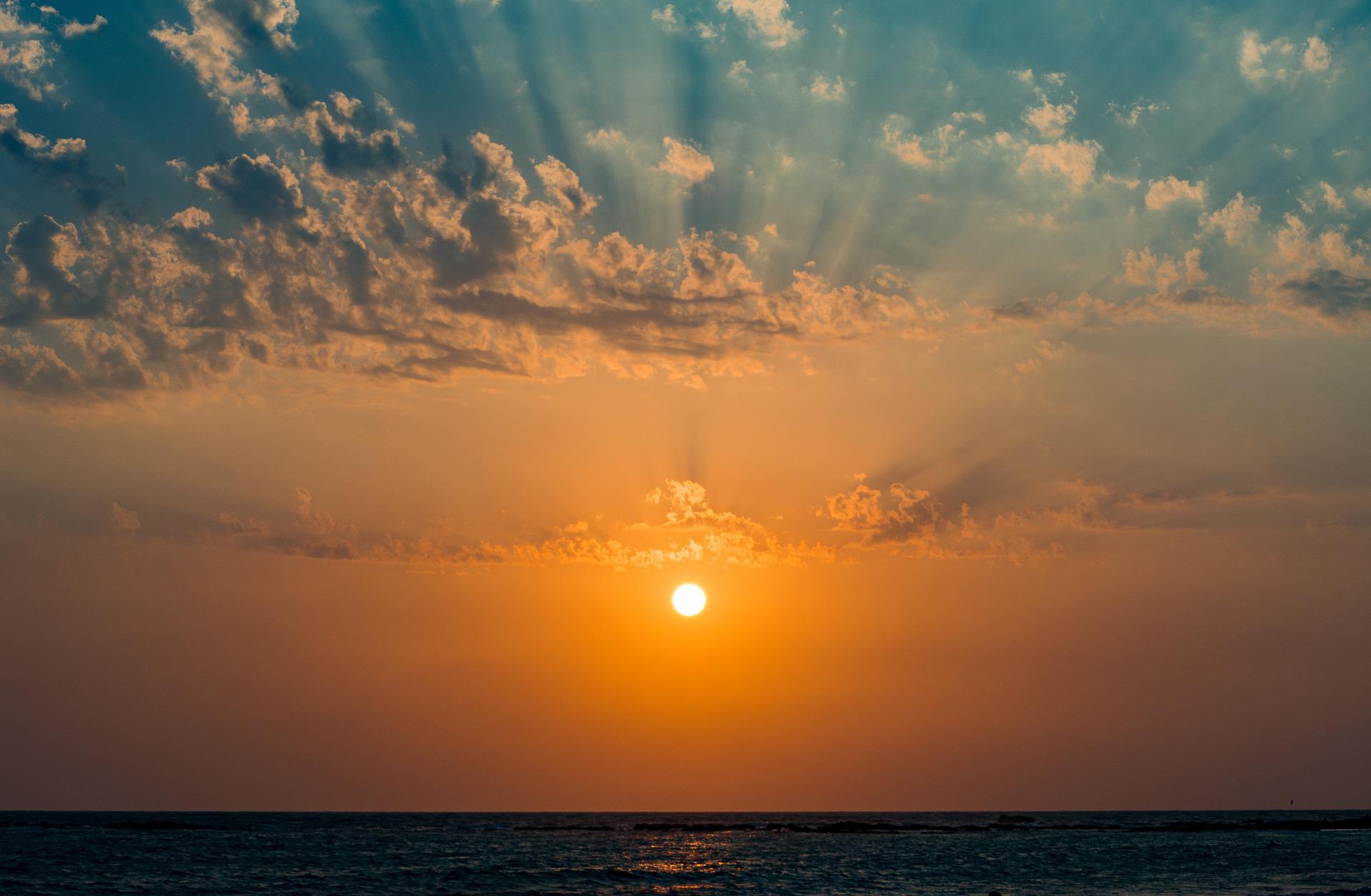 Above an ocean's horizon is an illuminating sun. Its ray spread forth into the beautiful light blue sky and its angelic clouds.