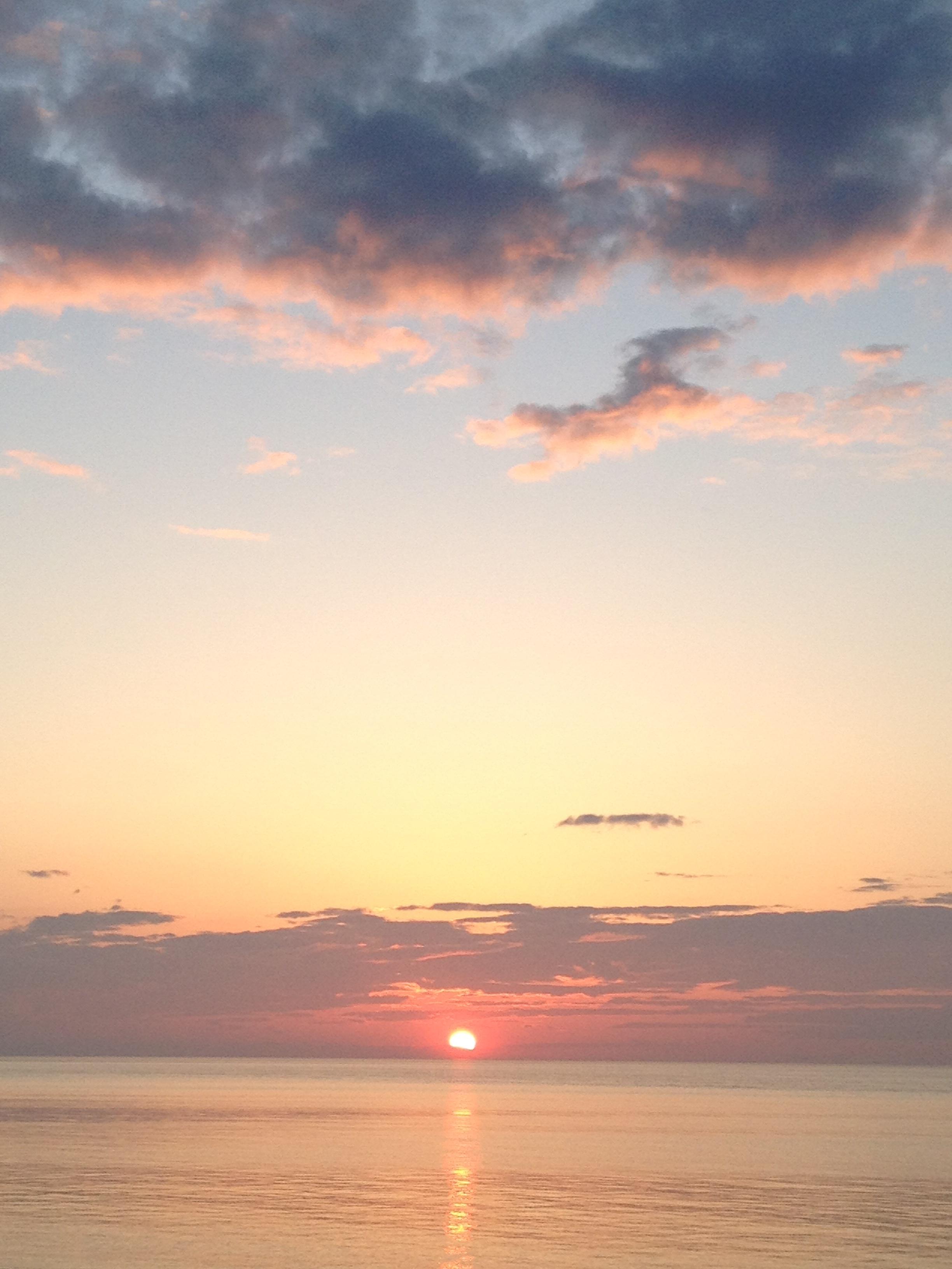 A photo of the rising sun. The photo has a portrait orientation, so it is taller than it is wider. The sun is emerging over the ocean's horizon. At the top of the photo are clouds of a blue-pink colour and fluffy. The clouds just above the rising sun are long and flat.