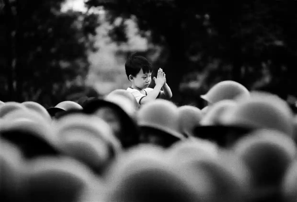 At Tiananmen Square, 1989. Just before the massacre. A small boy is on the shoulders of an adult, his hands in prayerful pose. All we see is the boy and many hard hats of Chinese soldiers.