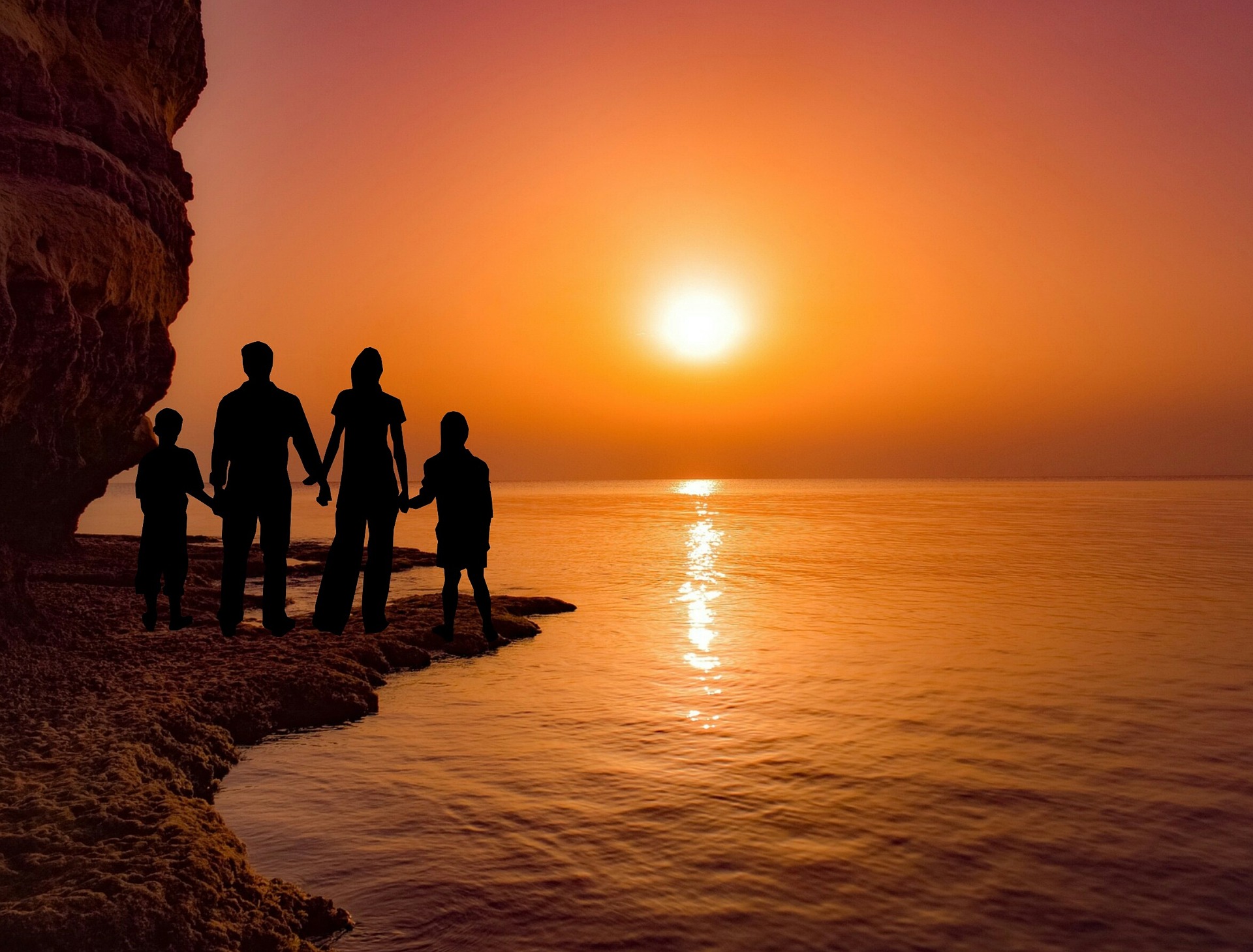 A sun sets over the sea casting an orange hue to the sky and sea. From a beach, by a giant rock, a silhouetted family of four looks on.