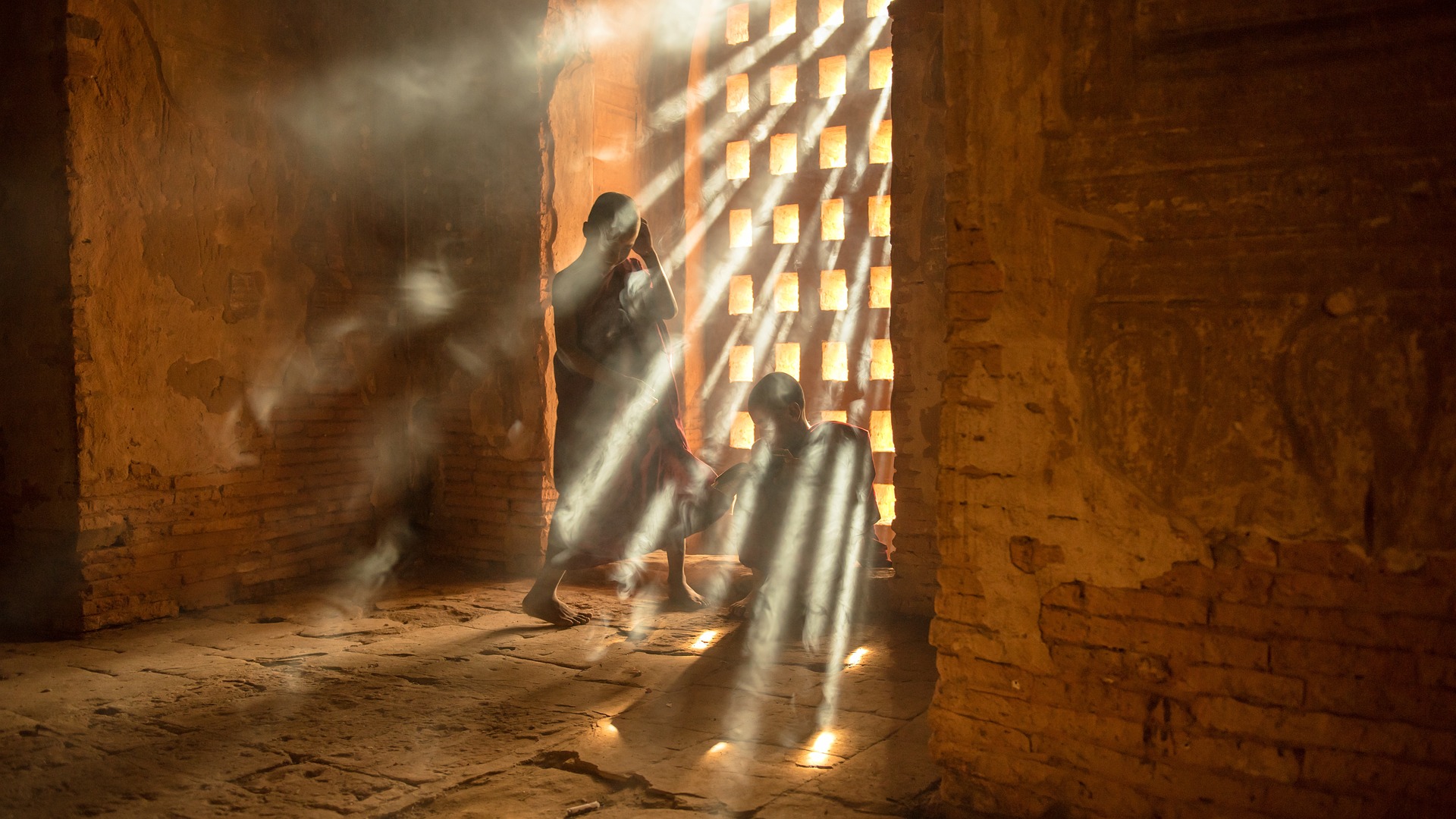 Two monk boys, one setaed, one standing, are in a dark room. They are by a wooden portcullis-style gate or window. Very strong sunrays pierce the openings and land on the dark interior.