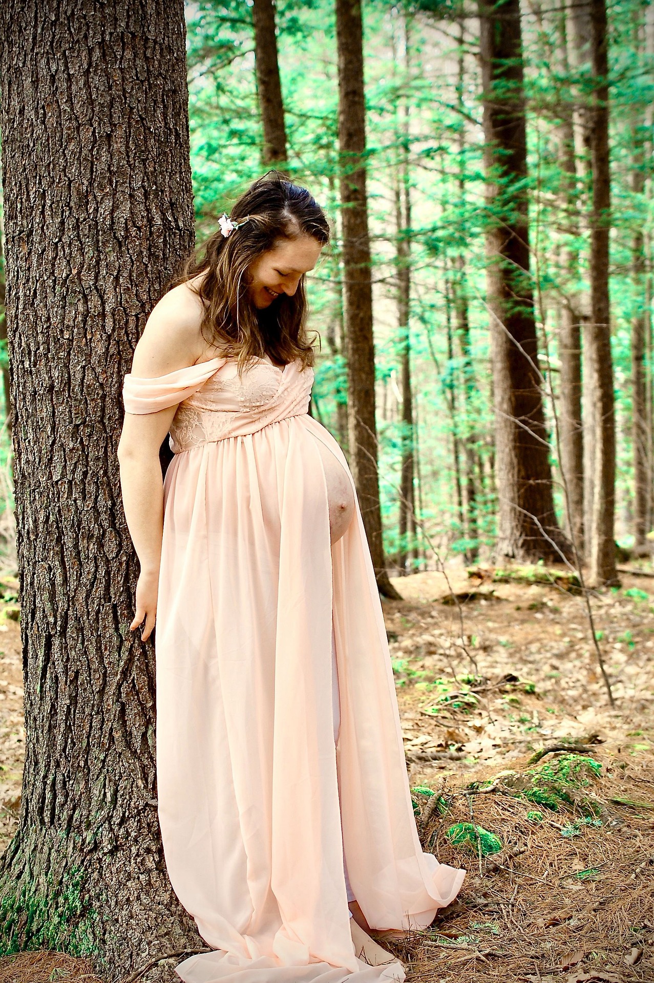 A pregnant woman stands with her back to a tree in a forest. She wears a Greek goddess robe. She looks down, greatly amused.