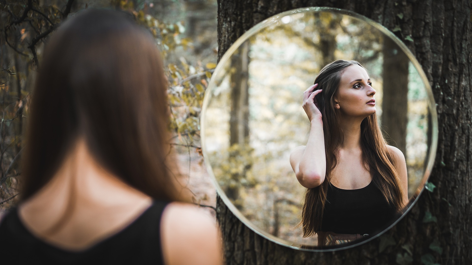 A young woman in a forest gazes at a round mirror on a tree. Her reflection doesn't look back at her; it looks off to the side, pensively stroking her hair.