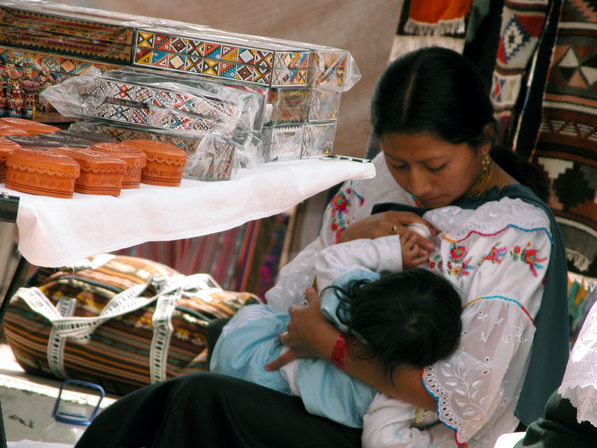 A woman, possibly indigenous from Ecuador, is seated and breastfeeding her toddler daughter. The mother seems to be at a market, selling indigenous crafts.