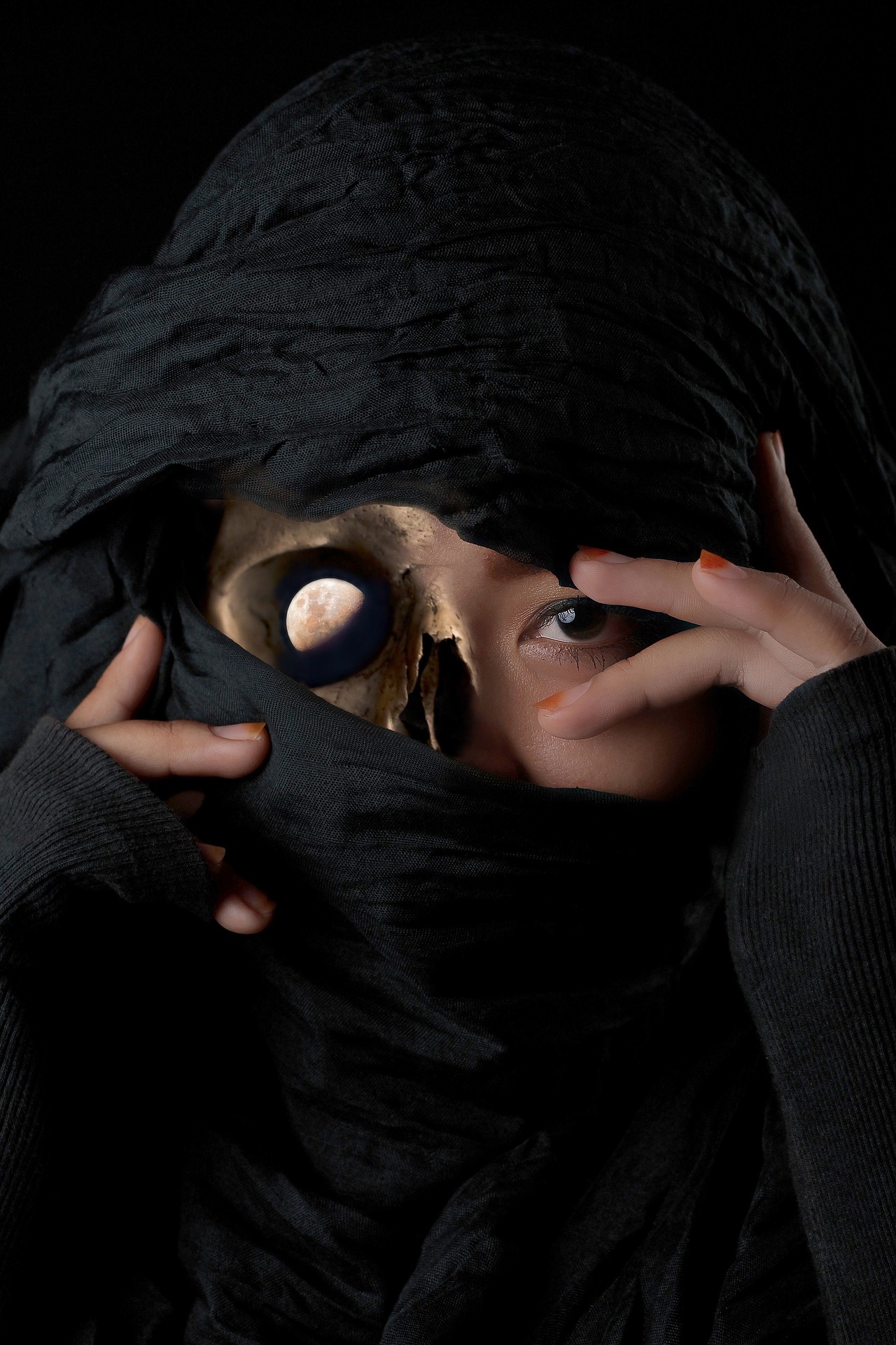 A ninja's head covered with a black wrapping. Mainly, her eyes are visible, one human, the other a planet. The nose is bone, as is a small part of her forehead. Her fingers with red nail polish frame her eyes.