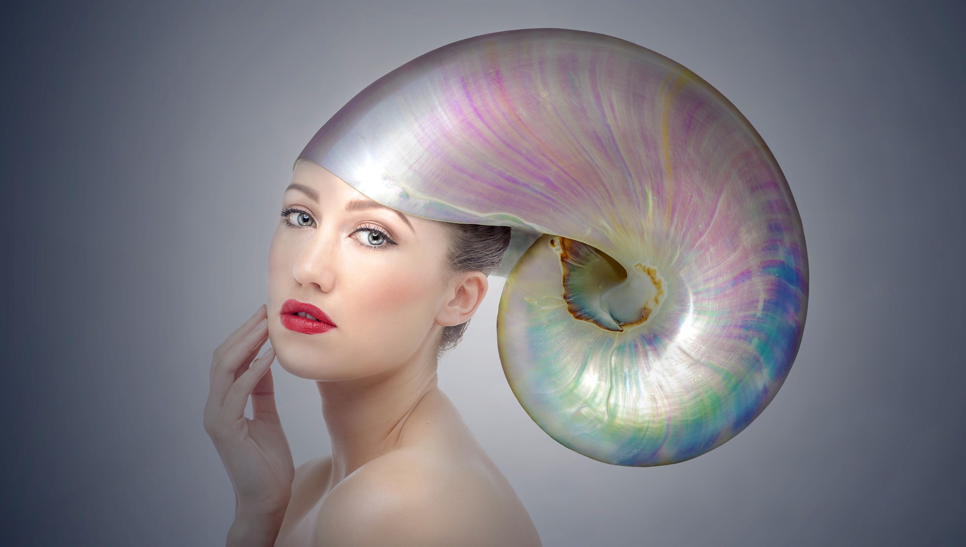 A woman with red lipstick looks at us, pensively. She wears a giant mother of pearl as a helmet. A pearl mother.