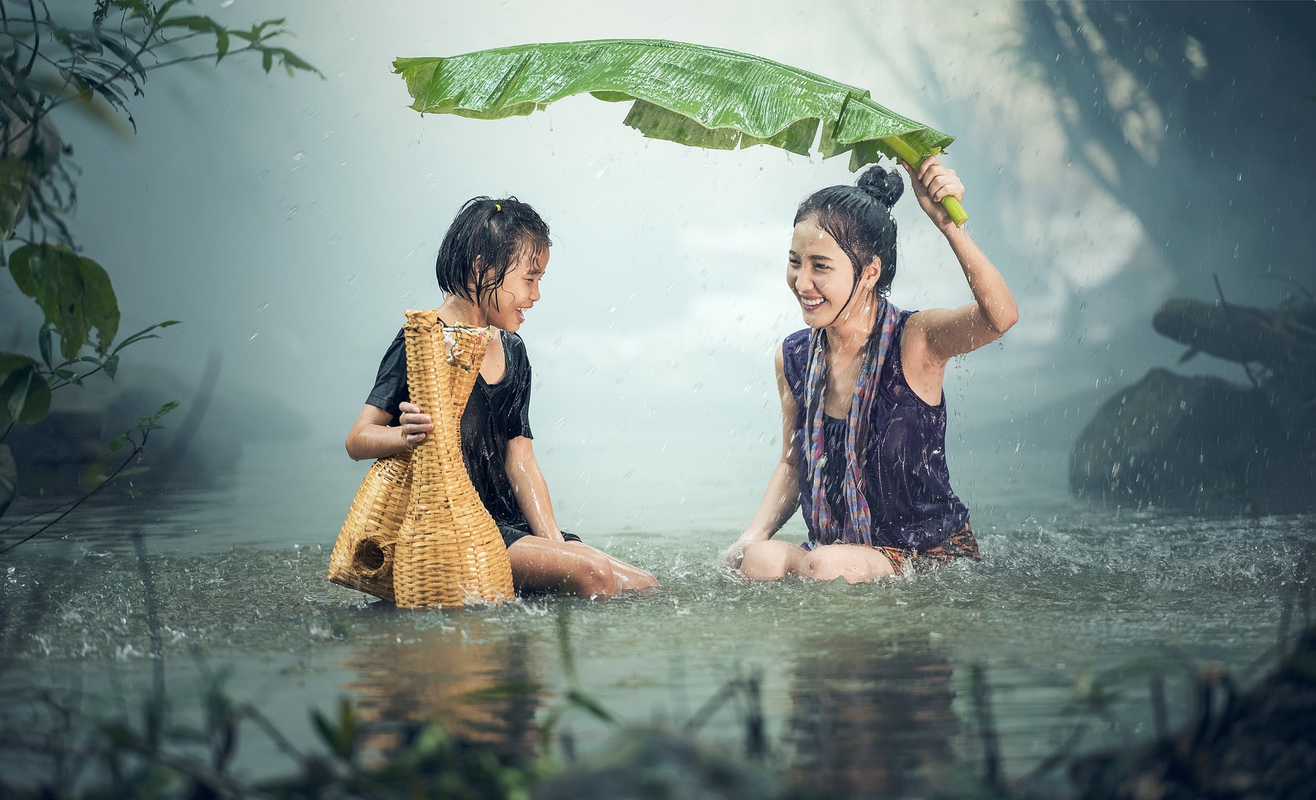 An  Asian mother and her daughter are in a tropical jungle pool. Their faces show great joy as the rain comes down and the Sun illuminates the scene with soft, bright light. The mother holds a large palm leaf to shield them. The daughter carries what appears to be two wicker containers.