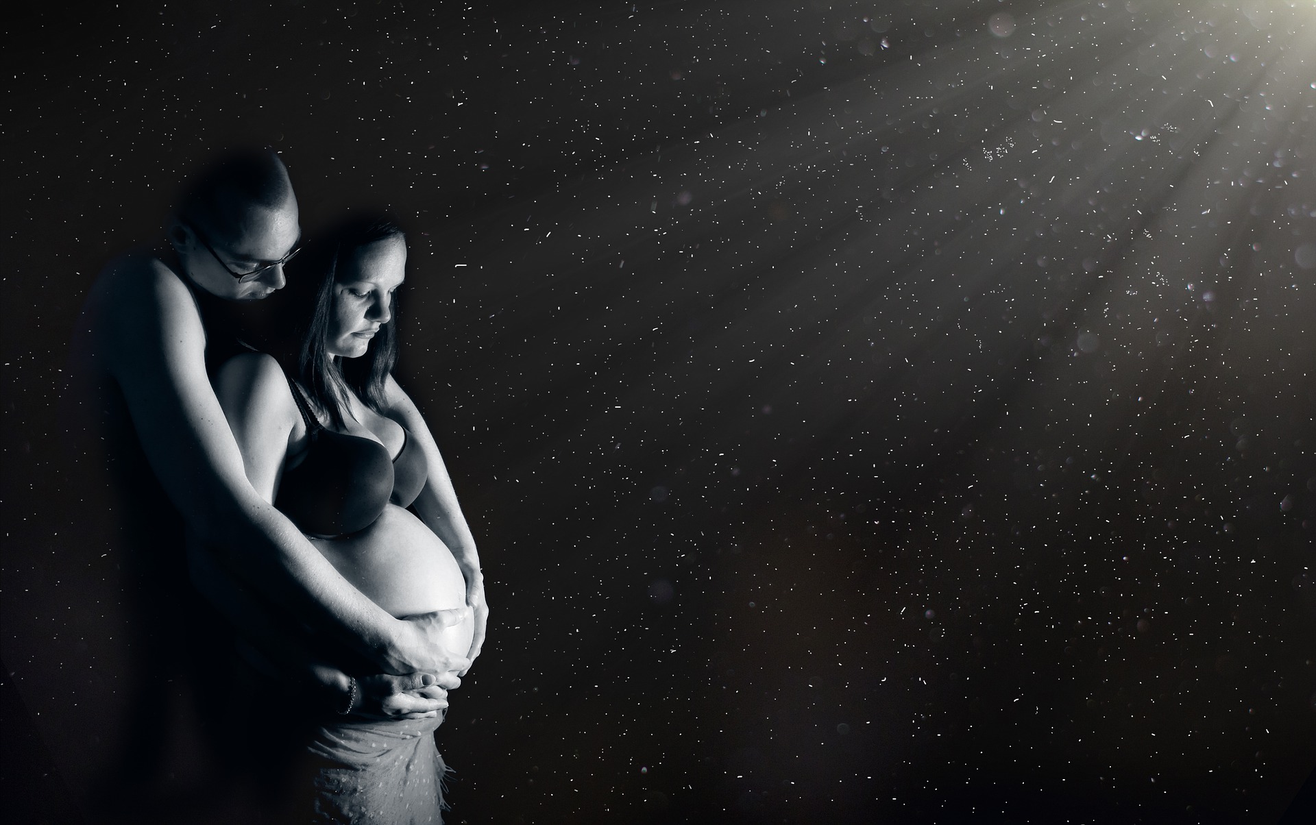 A pregnant woman is lovingly embraced from behind by her partner. The background is a starry sky with a brighter light source from the top right. It is all in black and white.