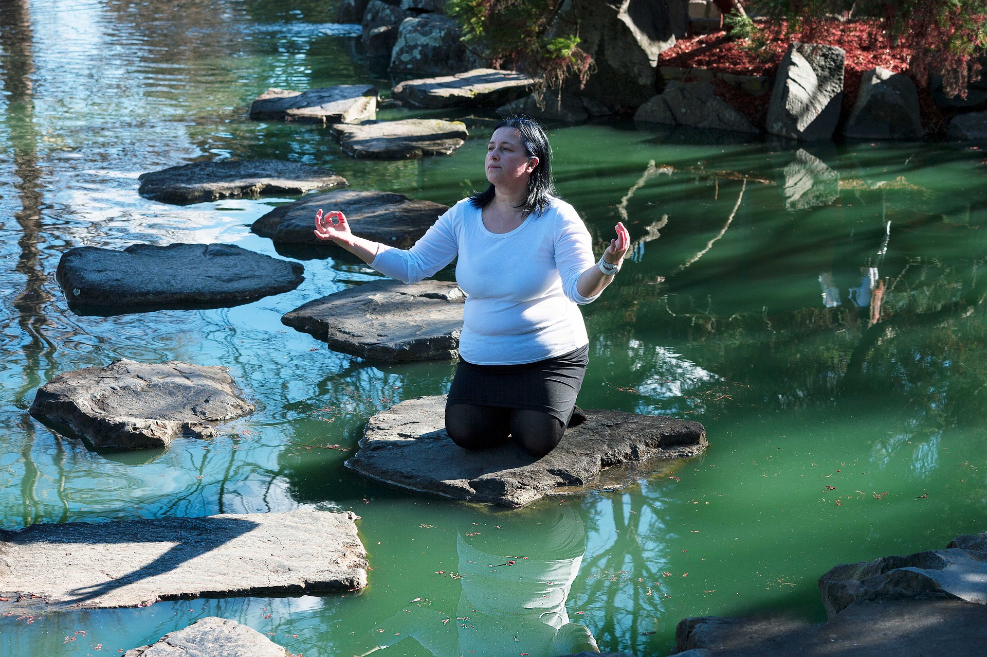 A beautiful pond with large stepping stones. A spiritual woman kneels on one of the stepping stones. Her hands are outstretched and she appears to be communing with the universe.