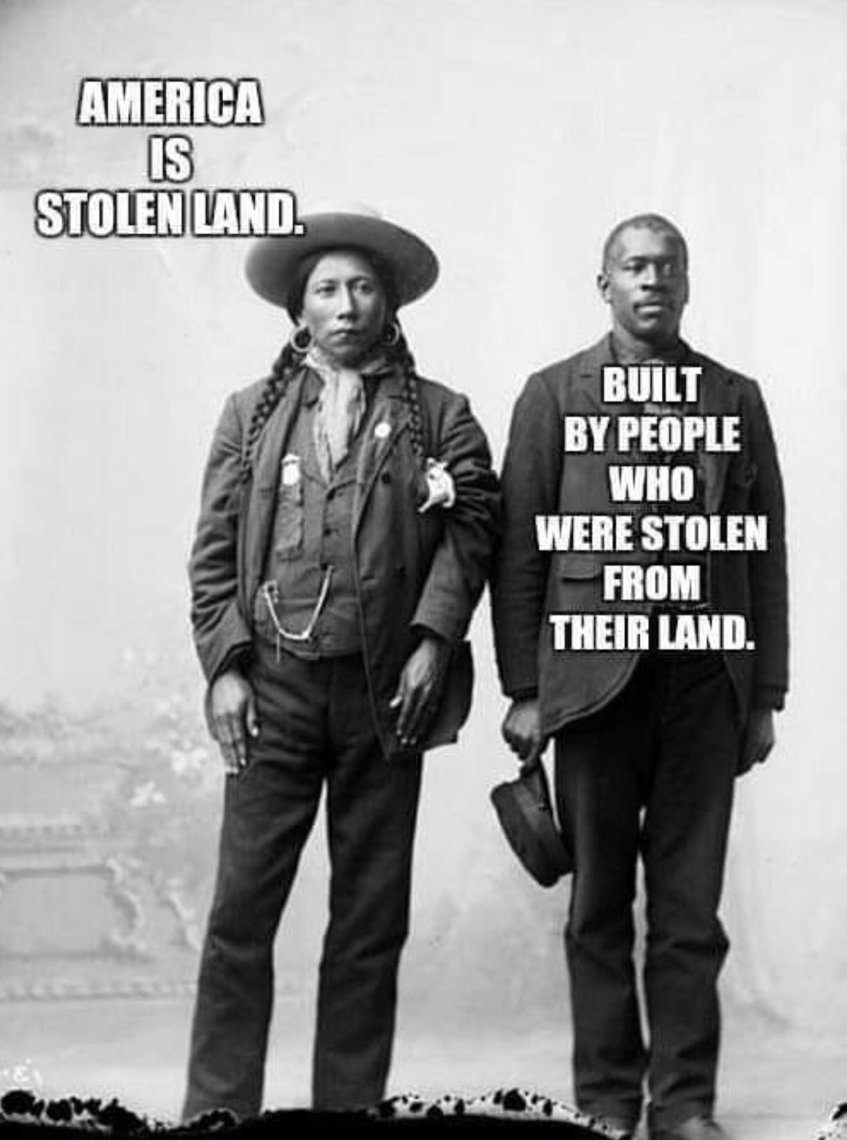 An old black-and-white photo. A Native American man stands next to a Black man. By the Native American man are the words 'America is stolen land'. By the Black man are the words 'Built by people who were stolen from their land'.