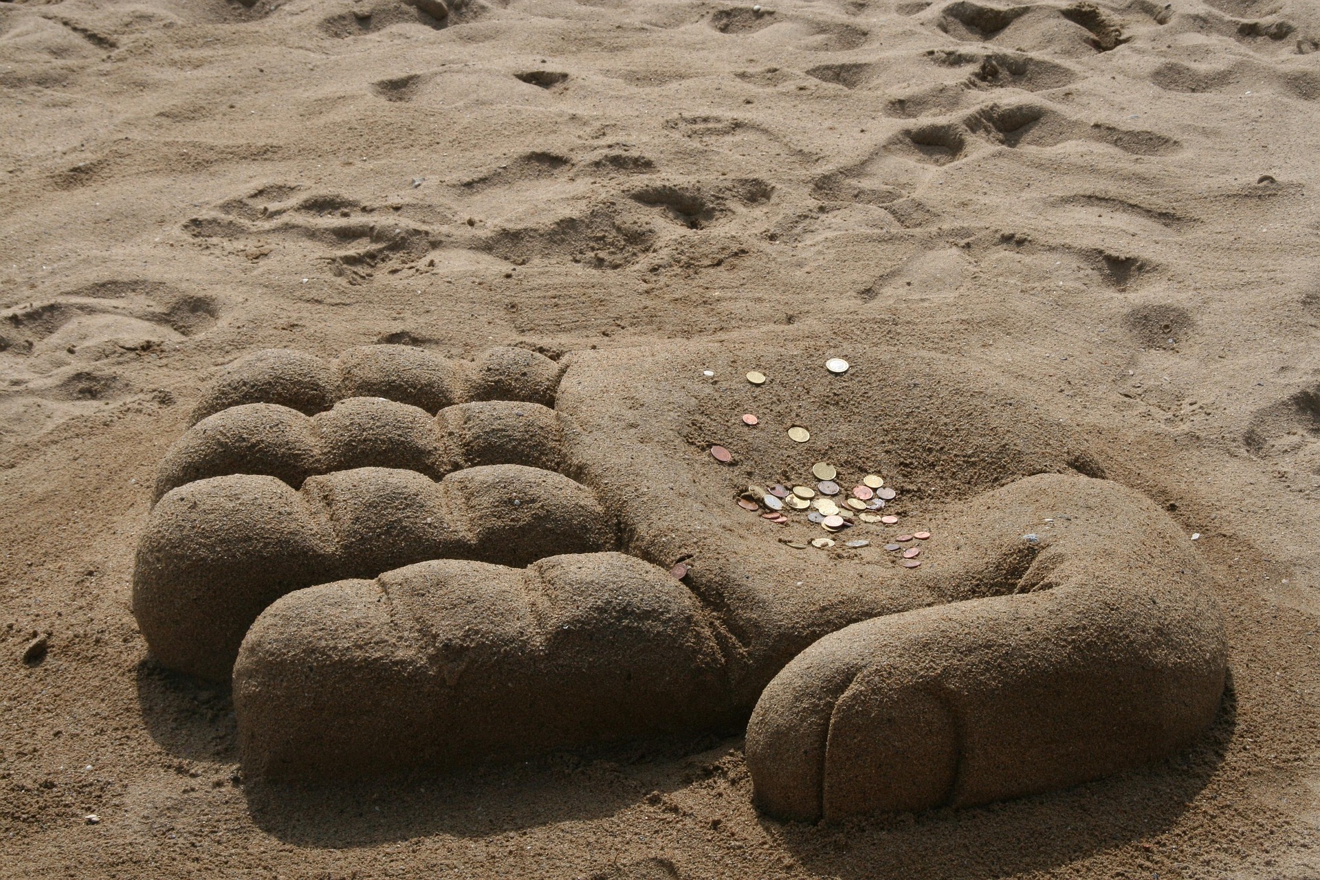On a beach, apparently a giant stone hand. It emerges from the sand, palm up. The palm has many coins. The giant stone sand is created from sand.