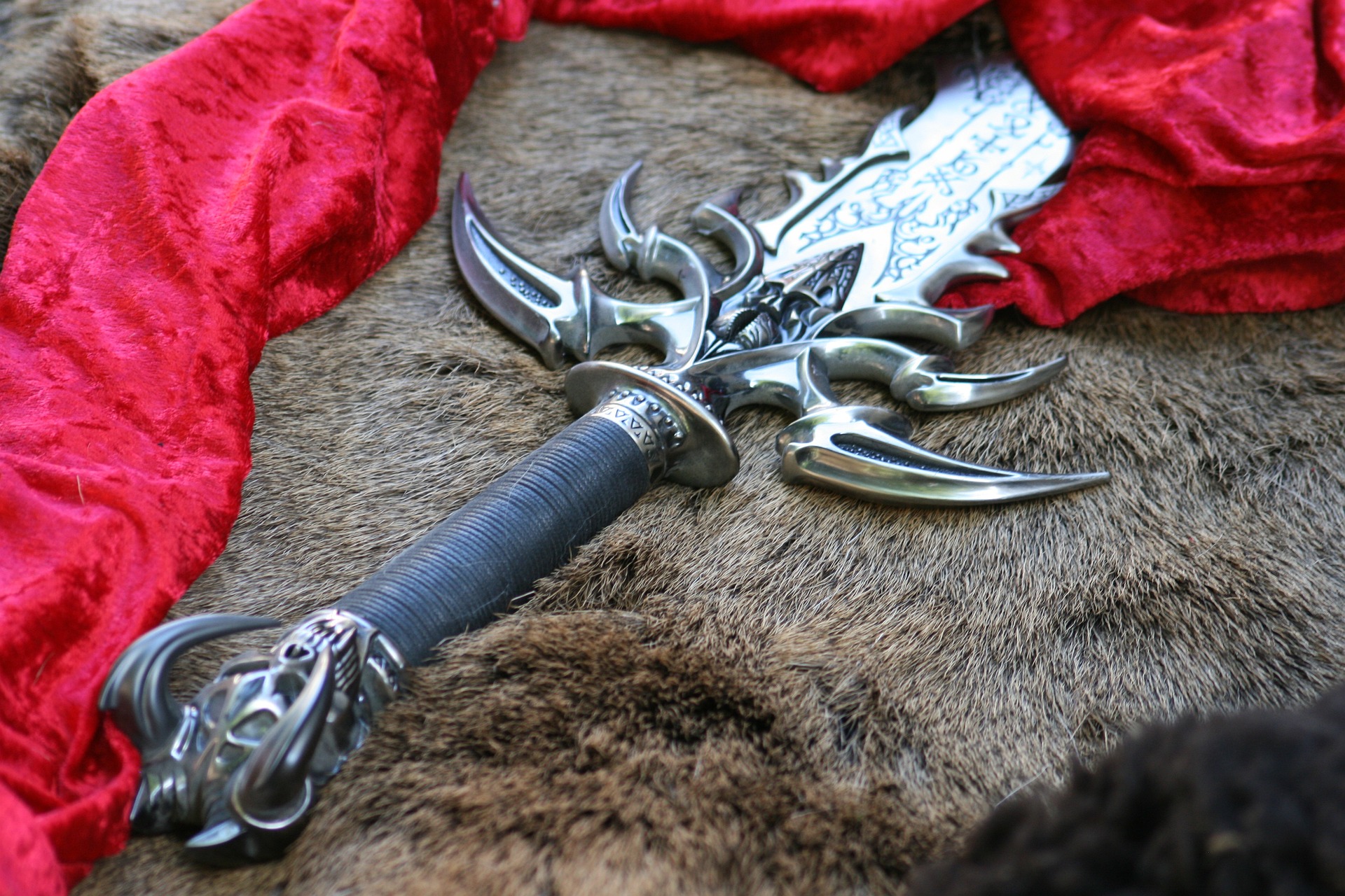Ornate sword. It has an intricate metal hilt that suggests fantasy, magic. A rich red cloth lies next to it.