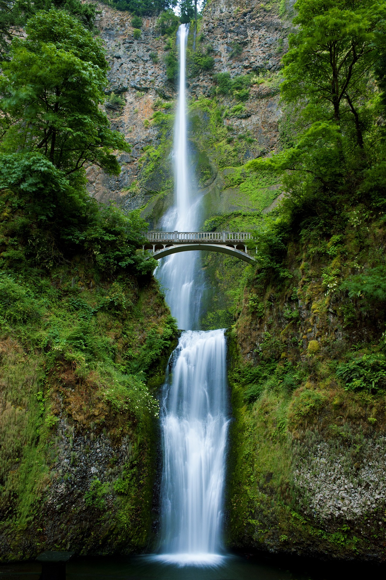 A long waterfall with greenery around. Halfway down, there is a wooden viewing bridge.