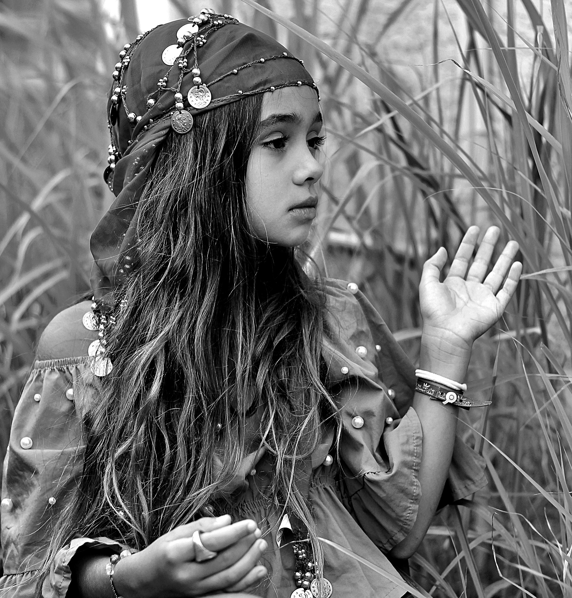 A black-and-white photo of a gypsy girl, sitting in nature, looking and gesturing to the right. She wears a scarf with 'coins' on her head.