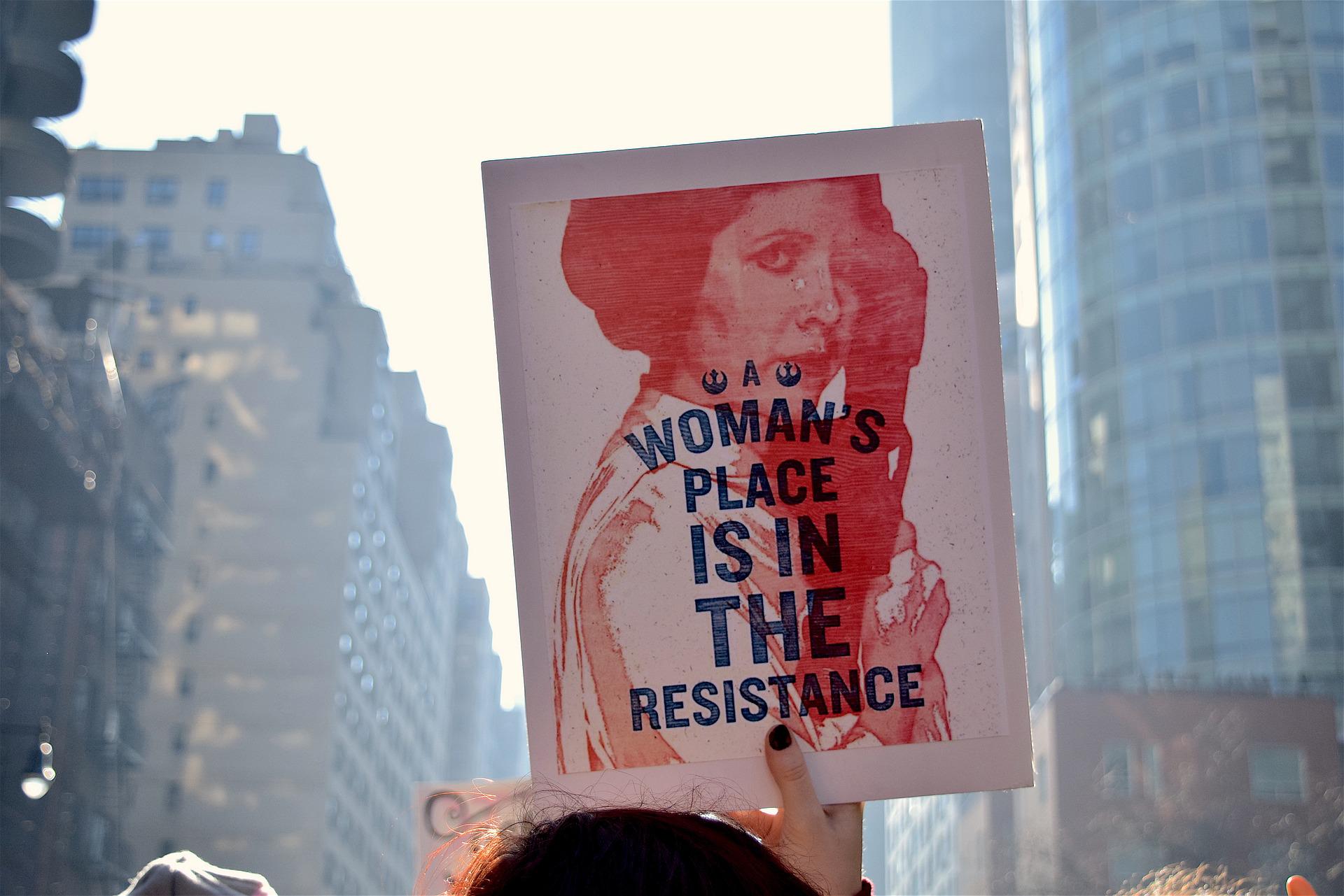 On a woman's march, on a street with skyscrapers, we see a protest placard held aloft. It shows a woman and the words 'A woman's place is in the resistance'.